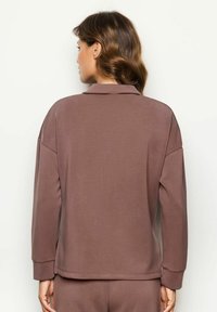 Mujer con cabello castaño ondulado que lleva una blusa suelta de manga larga en color malva y pantalones a juego, mostrada de espaldas contra un fondo liso.