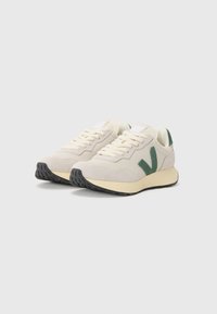 Ténis com um upper de malha bege claro, atacadores creme, detalhe do logo V em verde e uma sola de borracha preta texturizada. Design minimalista.