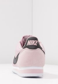 Nike-sneaker med en överdel i rosa mocka, mörkgrå detaljer, en vit mellansula och en framträdande svart logotyp på hälen. Klassisk design.