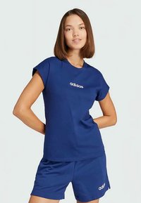 Tânără cu păr drept și brunet, purtând un tricou Adidas bleu și pantaloni scurți asortati, pozând cu mâinile în spate.