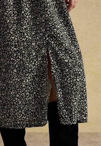 Schwarzes Stoffkleid mit weißen abstrakten Leopardenmustern, seitlichem Schlitz und weicher Textur. Jahr: ergänzt durch knielange schwarze Stiefel.