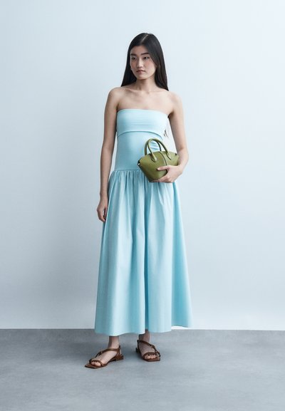 Νεαρή γυναίκα με αμανικό, μακρύ φόρεμα σε ανοιχτό γαλάζιο χρώμα, κρατώντας μια μικρή τσάντα σε ελιά πράσινο, με καφέ επίπεδα σανδάλια, στέκεται σε γκρι δάπεδο.