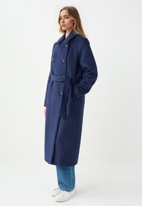 Marineblauwe dubbele-breasted jas met een brede kraag, riem en knoopaccenten. Gemaakt van wol, met een rechte, lange snit. Draagt over spijkerbroeken en sneakers.