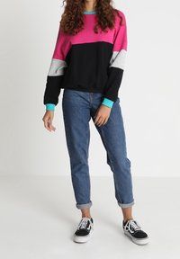 Sweat-shirt coloré avec des panneaux rose, gris et noir, avec un col et des poignets turquoise. Porté avec un jean en denim retroussé et des baskets.