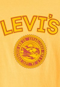 Sárga pamut póló, amelynek frontján feltűnő vörös betűkkel szerepel a "LEVI'S" felirat, valamint egy kör alakú grafika hegyekkel, nappal és az "ESTABLISHED 1873" szöveggel.