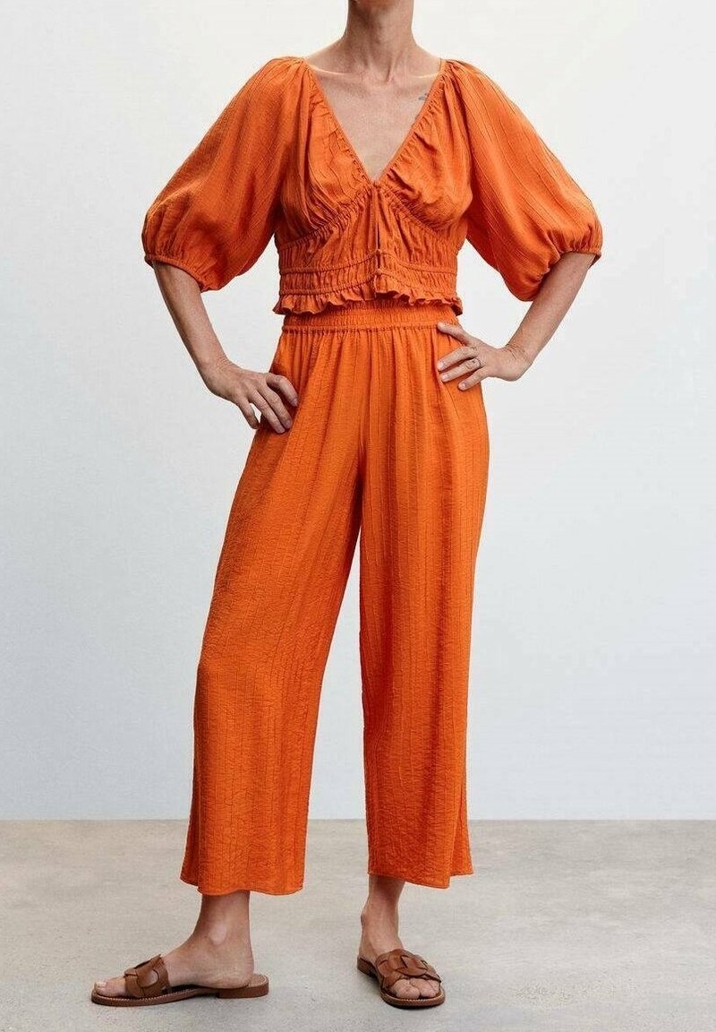 Oranžový, texturovaný dvoudílný outfit obsahující zkrácený top s nafouknutými rukávy a vysokými, volnými kalhotami. Zahrnuty ploché sandály.