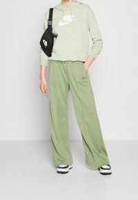 Ljust grön Nike-hoodie med en framficka, matchande gröna sweatpants och en svart axelremsväska med vita logotypdetaljer.