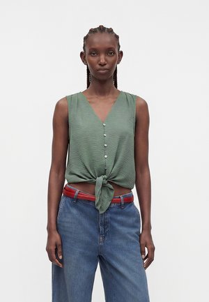 Kvinde med flettet hår iført ærmeløs grøn knapbluse bundet i taljen, rødt bælte og løse blå jeans, stående mod en ensfarvet baggrund.