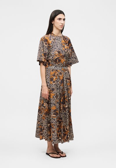 Γυναίκα στέκεται φορώντας midi φόρεμα με καφέ, μαύρο και μπεζ λουλουδάτο μοτίβο, με κοντά φαρδιά μανίκια και μαύρα σαγιονάρες.