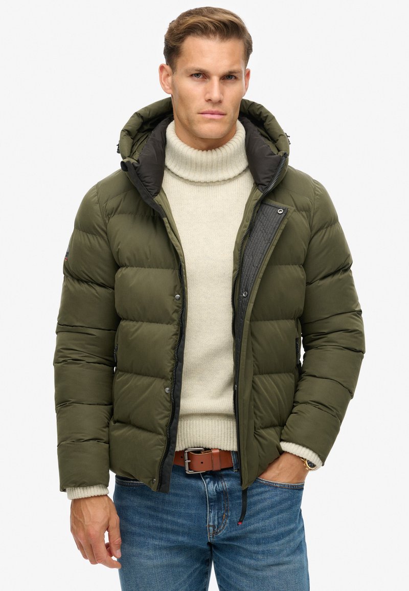 Chaqueta acolchada olive con cierre de cremallera y broches, capucha ajustable y ribete negro. Llevada sobre un suéter de cuello alto crema y jeans azules.