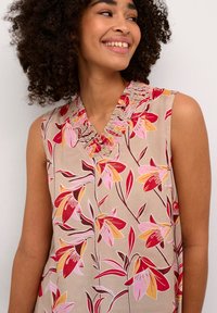 Bloemrijke mouwloze top in beige, met een geplooide, gestructureerde halslijn. Het ontwerp bevat rode en roze bloemen tegen een bladpatroon.