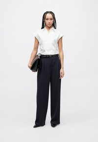 Λευκό πουκάμισο με κοντά μανίκια και μαύρο ψηλόμεσο παντελόνι. Το μοντέλο κρατάει ένα μαύρο φάκελο, με μινιμαλιστικό σχέδιο, λεία υφή και ραμμένη εφαρμογή.