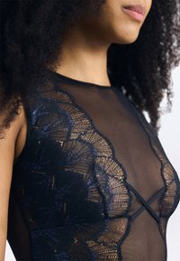 Juodas permatomas bodysuit'as, puošiamas sudėtingais gėlėtais nėriniais ir kryžminio dizaino priekyje. Textūruota medžiaga su lygiais kraštais.