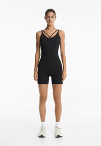 Zwart bodysuit met een aansluitend ontwerp, voorzien van gekruiste banden bij de halslijn en een soepele textuur. Gecombineerd met witte sneakers.