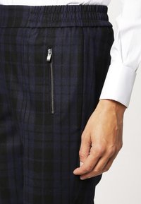 Granatowe spodnie w kratę z elastycznym pasem, z boczną kieszenią na zamek po lewej stronie. Materiał wydaje się miękki z delikatną teksturą.