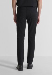 Persona vestida con pantalones negros ajustados y zapatos negros, de pie con la espalda hacia la cámara frente a un fondo claro y liso.