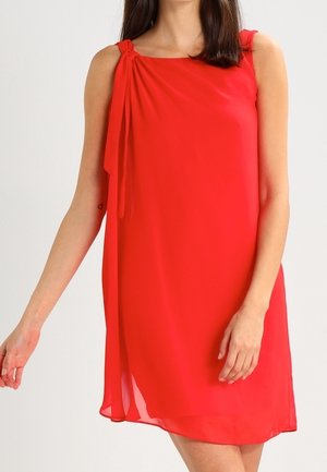 Femme portant une robe rouge sans manches avec un détail noué rassemblé sur l'épaule droite, debout devant un fond clair uni.