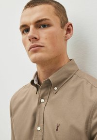 Camicia button-up marrone chiaro con colletto e logo di cervo ricamato sul petto. Presenta bottoni bianchi e una fodera a strisce a contrasto.