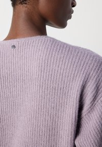 Ceriņkrāsas adīta džemperis ar ribota tekstūra, noapaļots kakla izgriezums un neliela metāla logo detaļa mugurpusē pie apkakles.