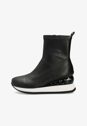 Botín negro de cuero hasta el tobillo con parte superior suave, entresuela blanca y un elegante detalle en cadena negra brillante en el tacón cuña, que presenta una suela texturizada.