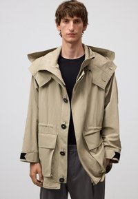 Beige huva jacka tillverkad av lätt tyg, med dragkedja framtill, stora fickor och knappdetaljer på ärmarna.