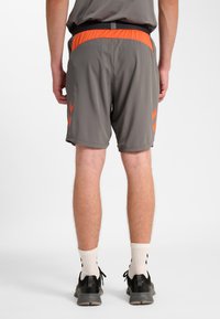 Gråa herrsportshorts med orange accenter, utrustade med ett midjeband i mesh och chevronmönster på sidorna. Lättviktigt, andningsbart tyg.