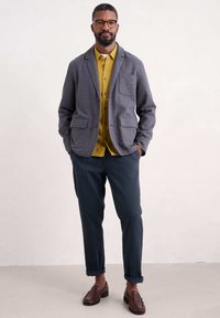 Blazer a scacchi blu, camicia gialla con bottoni, pantaloni scuri arrotolati e scarpe slip-on intrecciate marroni. Design casual con diverse tasche.