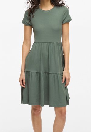 Femme portant une robe vert olive ample, courte, à manches courtes, avec une jupe à volants et un col rond, debout devant un fond uni.