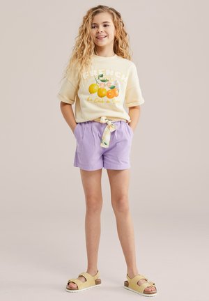 Osmijeh djevojke s dugom kovrčavom kosom nosi krem košulju s printom voća, ljubičaste kratke hlače s vezanim remenom i žute sandale otvorenih prstiju.