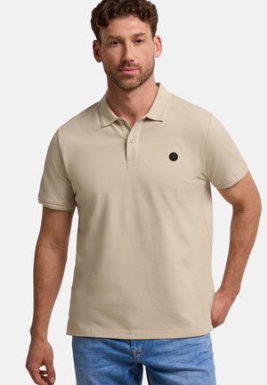 Homme aux cheveux courts et bouclés, portant un polo beige à manches courtes et un jean bleu, debout avec une main dans la poche.
