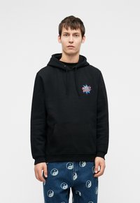 Fiatal férfi fekete kapucnis pulcsiban, rajta a "Santa Cruz" logóval és kék farmerben, amelyet fehér yin-yang szimbólumok mintáznak, egy egyszínű háttér előtt áll.