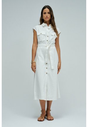 Frau steht und trägt ein ärmelloses weißes Midikleid mit durchgehender Knopfleiste, einem Taillengürtel und braunen Riemchensandalen vor einem schlichten Hintergrund.