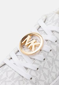 Zapatilla blanca con un patrón texturizado, que presenta un destacado acento del logo dorado de MK y cordones blancos.