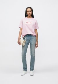 Hellrosa Baumwoll-T-Shirt mit weißem und rotem Logo, kombiniert mit hellblauen Jeans und weißen Sneakers. Model hält eine beige Handtasche.