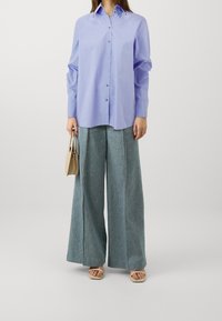 Camicia button-up viola chiaro con maniche lunghe, abbinata a pantaloni in denim a gamba larga di una sfumatura di blu chiaro. Tiene una piccola borsa beige.