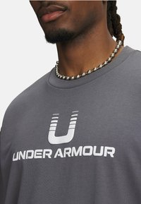 Grå kortærmet skjorte med "UNDER ARMOUR" logo i hvid. Materialet er glat, og der er en rund halsudskæringen. Sølv perlekæde synligt.