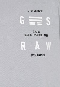 Camiseta de algodón gris con texto en blanco y negro en negrita: "G-STAR RAW" y "JUST THE PRODUCT 1989." Diseño simple y casual.