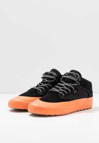 Schwarze High-Top-Sneaker mit orangefarbener Gummisohle, grauen Schnürsenkeln und Wildledertextur. Sie haben eine abgerundete Zehenpartie und ein dezentes Logodetail.