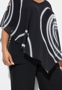 Poncho in chiffon nero con motivi circolari bianchi, caratterizzato da una forma rilassata e un design a strati, indossato sopra un top e pantaloni neri.