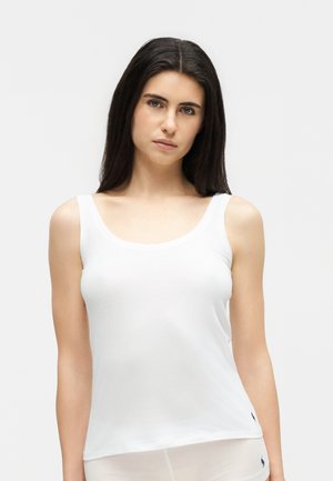 Γυναίκα με μακριά σκούρα μαλλιά που φοράει λευκό αμάνικο τοπ σε απλό φωτεινό φόντο, κοιτώντας απευθείας την κάμερα.