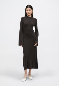 Brązowa dzianinowa sukienka z długim rękawem, stójką, dopasowanym gorsetem i szerokimi rękawami, zestawiona z białymi mule i dużymi kolczykami w kształcie kół.