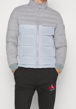 Mand iført en grå og lyseblå vatteret jakke med lynlåslommer og sorte bukser med rød "BOSS" tekst og en lille grafik.