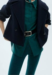 Cappotto doppiopetto blu navy, blusa color teal, cintura nera e pantaloni teal con texture. Finiture in tessuto morbido con colori a contrasto e bottoni lucidi.