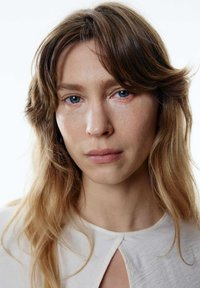 Porträt einer Person mit hellbraunen, gewellten Haaren, die ein strukturiertes, cremefarbenes Kleidungsstück mit einem Schlüsselloch-Ausschnitt trägt, und natürlichem Make-up.