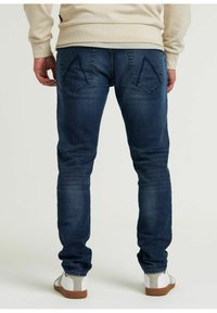 Jeans in denim blu scuro con una vestibilità slim, dotati di tasche posteriori inclinate e dettagli cuciti, abbinati a scarpe casual di colore chiaro.