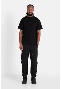 Zwarte katoenen t-shirt met een klein rood logo, gecombineerd met zwarte cargobroeken met meerdere zakken en een gestructureerde stof. Zwarte sneakers.