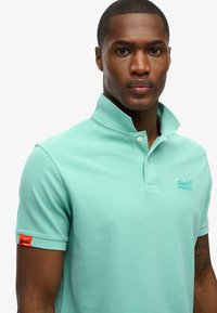 Polo shirt turquoise clair en tissu texturé, avec un col montant et trois boutons. Inclus un patch de logo orange contrastant.