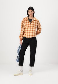 Oranžinės ir grietinėlės spalvos patalynės megztinis, su pusiau užtrauktuku, ilgomis rankovėmis ir juodomis kelnėmis. Modelis laiko mėlyną maišą ir užsideda baltus sportbačius.