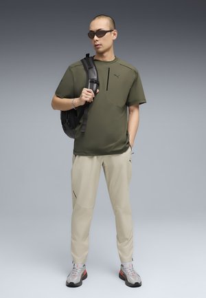 Hombre con gafas de sol, camiseta verde ejército de Puma, pantalones beige y zapatillas, lleva una mochila negra en un hombro, de pie sobre un fondo gris.