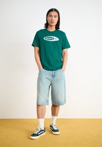 Žalias medvilnės marškinėlis su baltu Reebok logotipu, derinamas su šviesiai mėlynomis džinsinėmis šortais ir tamsiai žaliais sportbačiais su baltais akcentais.
